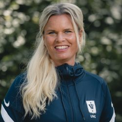 En smilende Andrine Holtan med langt blondt hår iført en sporty jakke med logo på, stående utendørs med uskarp grøntbakgrunn.