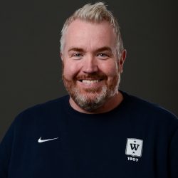En smilende mann, Espen Fossli Olaussen, med kort skjegg og lyst hår, iført mørk Nike-sportsskjorte med et merke som inkluderer initialene "W" og