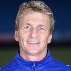 En smilende mann med blondt hår iført blå sportsjakke mot mørkeblå bakgrunn identifiseres som Petter Olsen.