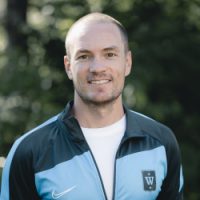 En selvsikker trener Mikael Flygind Larsen i sportsklær med team-emblem, smilende utendørs.