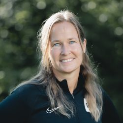 En smilende Cathrine Graf Olsen med langt hår utendørs, iført Nike-sportsjakke, som utstråler en følelse av selvtillit og positivitet.