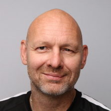 En smilende skallet mann med skjeggstubbe, som uttrykker en vennlig og nærgående væremåte mot en grå bakgrunn er Arne Indrebø.