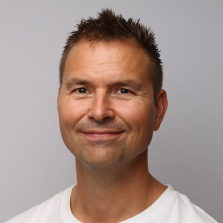 En mann med et fornøyd smil, kort hår og en lys skjeggstubbe, iført en hvit t-skjorte med rund hals mot nøytral bakgrunn er Atle Sigmundsen.