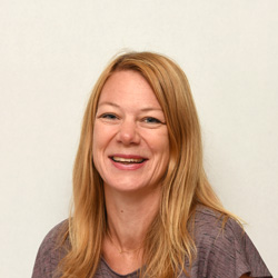 En smilende kvinne med langt rett blondt hår, iført en Camilla Horne Heidum brun topp, satt mot en ensfarget hvit bakgrunn.