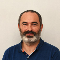 En mann med kort skjegg og bart som smiler til kameraet, iført en blå poloskjorte med oransje detaljer er Cyril Hébert.