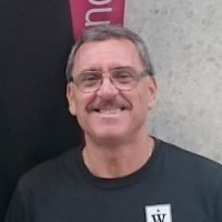 En smilende Dan Simion med briller iført en svart t-skjorte, stående foran en grå teksturert vegg med et rosa og hvitt skilt over hodet.