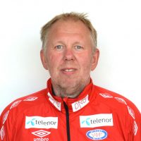 Portrett av Gjermund Østby i rød sportsjakke med sponsorlogoer, smilende mot en vanlig hvit bakgrunn.
