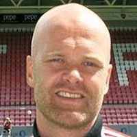 Jan Tore Ophaug, en mann med barbert hode, smiler til kameraet med en stadionsittegruppe i bakgrunnen.