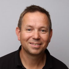 En smilende, middelaldrende mann med kort hår mot en nøytral grå bakgrunn er Kjell Skartveit.