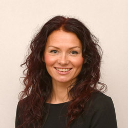 En smilende kvinne med langt krøllet kastanjebrunt hår mot en nøytral bakgrunn, som minner om Line Koppergårds stil.