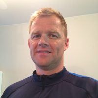 En mann med et selvsikkert smil iført en blå og svart atletisk topp med glidelås, stående innendørs, er Martin Gjerde Lester.
