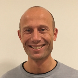 En smilende Martin Strømner med barbert hode, iført grå t-skjorte mot nøytral bakgrunn.