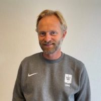 En smilende mann med skjegg, iført en grå nike sportsskjorte med logo på høyre bryst, identifisert som Morten Brevik, står foran en vanlig vegg.