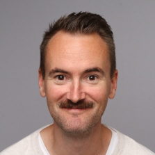 En blid Olav Veland Thu med kort skjegg og bart, smilende til kamera mot en grå bakgrunn.