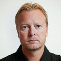 Et portrett av Petter S. Wilhelmsen med kort blondt hår, intenst blikk og svart skjorte mot ensfarget bakgrunn.