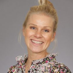 En glad kvinne med et strålende smil, blond toppknutefrisyre, iført en blomstermønstret bluse designet av Siv Askheim, mot en nøytral grå bakgrunn.