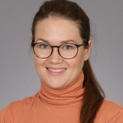 En smilende kvinne med briller iført oransje turtleneck mot en grå bakgrunn, kjent som Marit Damhaug.