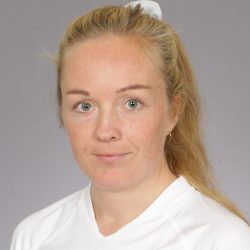 En kvinne, Anne Jorunn Tysseland, med lyst hår bakbundet, klare blå øyne, og et nøytralt uttrykk mot en grå bakgrunn.