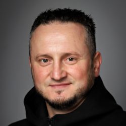 Portrett av Andriy Balyasnyy, en smilende mann med kort hårklipp og en svart glidelåstopp mot en grå bakgrunn.