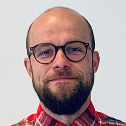 Portrett av en smilende mann med skjegg og briller iført en rød rutete skjorte.