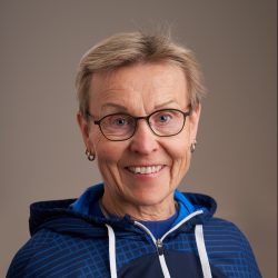 En blid seniorkvinne, Birgit Korvald, iført briller, med et varmt smil, kledd i en blå og svart atletisk jakke, som utstråler positivitet og aktive livsstilsvibber.