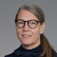 Et portrett av Gitte Madsen-Kaarød med skulderlangt hår, iført briller med rund innfatning og mørk rullekrave, smilende subtilt mot en grå bakgrunn.