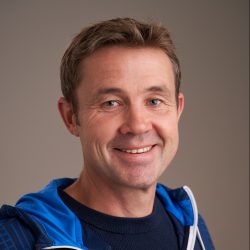 En smilende Harald Lindal med et varmt uttrykk, iført blå sportsjakke med hvite detaljer, satt mot en nøytral grå bakgrunn.