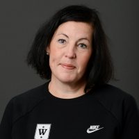 Portrett av Jenny Ervik, en kvinne med skulderlangt mørkt hår iført en Nike-genser med logo foran, satt mot en grå bakgrunn. Hun virker kontemplativ og har et subtilt smil