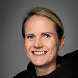 En kvinne med et vennlig smil, lys hud og kort blondt hår, iført svart topp, er Kirsti Damsgård Langeland mot en grå bakgrunn.