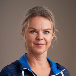 En selvsikker middelaldrende kvinne, Kjerstin Aasland Lae, med et mildt smil, iført blå sportsjakke mot nøytral bakgrunn.