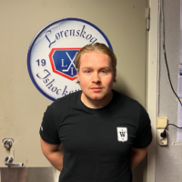 En person med langt blondt hår står foran en dør prydet med en «Lørenskog Ishockey»-logo. De har på seg en svart T-skjorte med et lite hvitt emblem på brystet. Døren og veggene er lyse, med noen papirer festet på høyre side. Denne personen er Mathias Falseth.