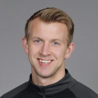En smilende Petter Seljeskog med kort blondt hår, blå øyne og en svart glidelåstopp mot grå bakgrunn.