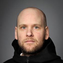 Scott Harrington, en mann med skallet hode og skjegg iført en svart hettegenser, ser alvorlig på kameraet mot en grå bakgrunn.