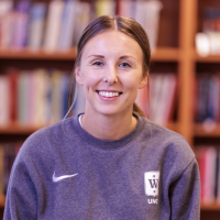 En smilende kvinne i en grå Nike-genser med en Oda Kvam Korbi-universitetslogo, stående foran bokhyller i en bibliotekomgivelse.