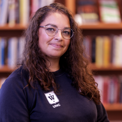 En smilende kvinne med krøllete hår iført briller og en svart t-skjorte med "Daniela Paz Silva"-logoen, stående foran en bokhylle fylt med bøker.