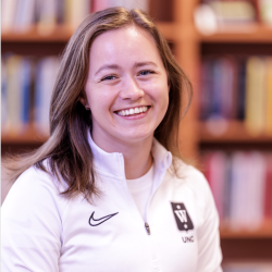 En munter ung kvinne, ved navn Sofie Bråten, med et strålende smil iført en hvit Nike-sportsjakke, poserer foran en uskarp bakgrunn av bokhyller, sannsynligvis i et bibliotek eller