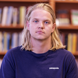 Ørjan Gabrielsen, en ung mann med skulderlangt blondt hår iført en blå Patagonia t-skjorte, står foran en bokhylle fylt med bøker og ser direkte på kameraet