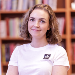 Selvsikker kvinne med et subtilt smil, iført en uformell hvit t-skjorte, i et bibliotek med hyller med bøker i bakgrunnen. Fotografi av Emilie Bergei.