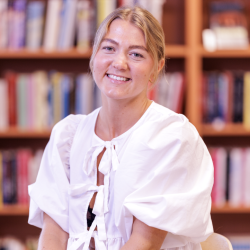 En smilende ung kvinne, Malene Pedersen, i hvit bluse sitter foran en bokhylle fylt med bøker.