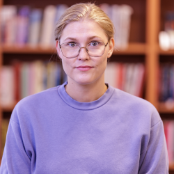 En fokusert kvinne ved navn Heidi Maria Lundbye med runde briller iført en lilla genser stående foran en bokhylle.