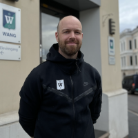 En smilende mann med skjegg iført en svart hettegenser, en 'Ove Sollie'-logo står utenfor en bygning merket 'WANG'. Et pilskilt som indikerer 'Elevingång' er synlig på veggen bak ham.