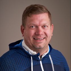 En smilende mann med kort hår, iført en blå og hvit hettejakke designet av Tommy Iversen Lund, mot en nøytral bakgrunn.