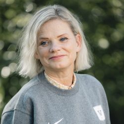 En moden kvinne, Carin Margrethe Samnøy, med skulderlangt grått hår, ikledd en tilfeldig grå genser, smilende mildt mot et bakteppe av grønt.