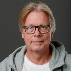Selvsikker middelaldrende mann, Arild Dragesæt, med briller og en uformell hettegenser poserer for et studioportrett mot en grå bakgrunn.