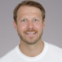 En smilende mann med lyse skjeggstubber og blå øyne, iført hvit t-skjorte mot grå bakgrunn, heter Joachim Eltvik.