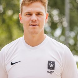 En ung idrettsutøver, Andreas Ringlund Hansen, i en hvit sportsskjorte med logo, poserer selvsikkert for et portrett utendørs med trær mykt uskarpe i bakgrunnen.