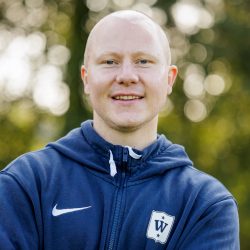 Eirik Pedersen, med et smil, iført blå sportsjakke og treningsklokke, stående utendørs med trær og bokeh-lys i bakgrunnen.