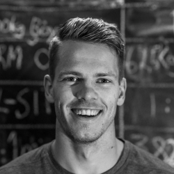 En ung mann med et muntert smil i svart-hvitt fotografi, som står foran en tavle med håndskrevet tekst og tall er Jonas Gudmundsen Lund.