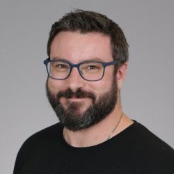 En smilende mann, Joshua Skidmore-Hornby, med skjegg og blåinnrammede briller, iført en svart skjorte mot en grå bakgrunn.
