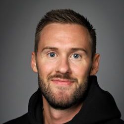 Selvsikker mann med kort skjegg og et hyggelig smil iført svart hettegenser på grå bakgrunn, identifisert som Kristian Søraker.